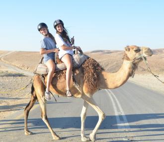 Two teens riding a camel across a street in the desert